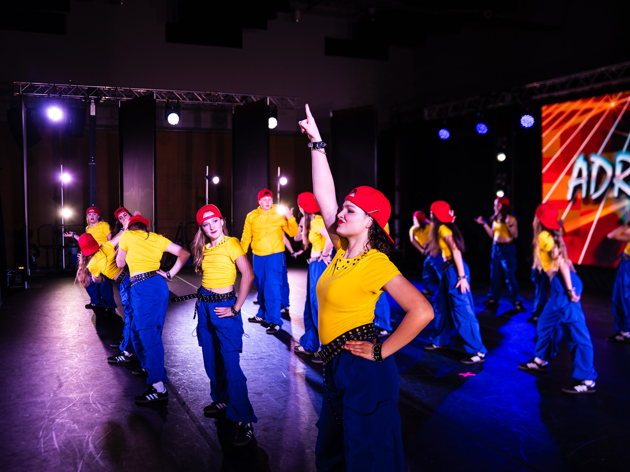 Dancer in the foreground with arm raised performs on stage; group of dancers in yellow shirts, blue pants, and red caps behind her.