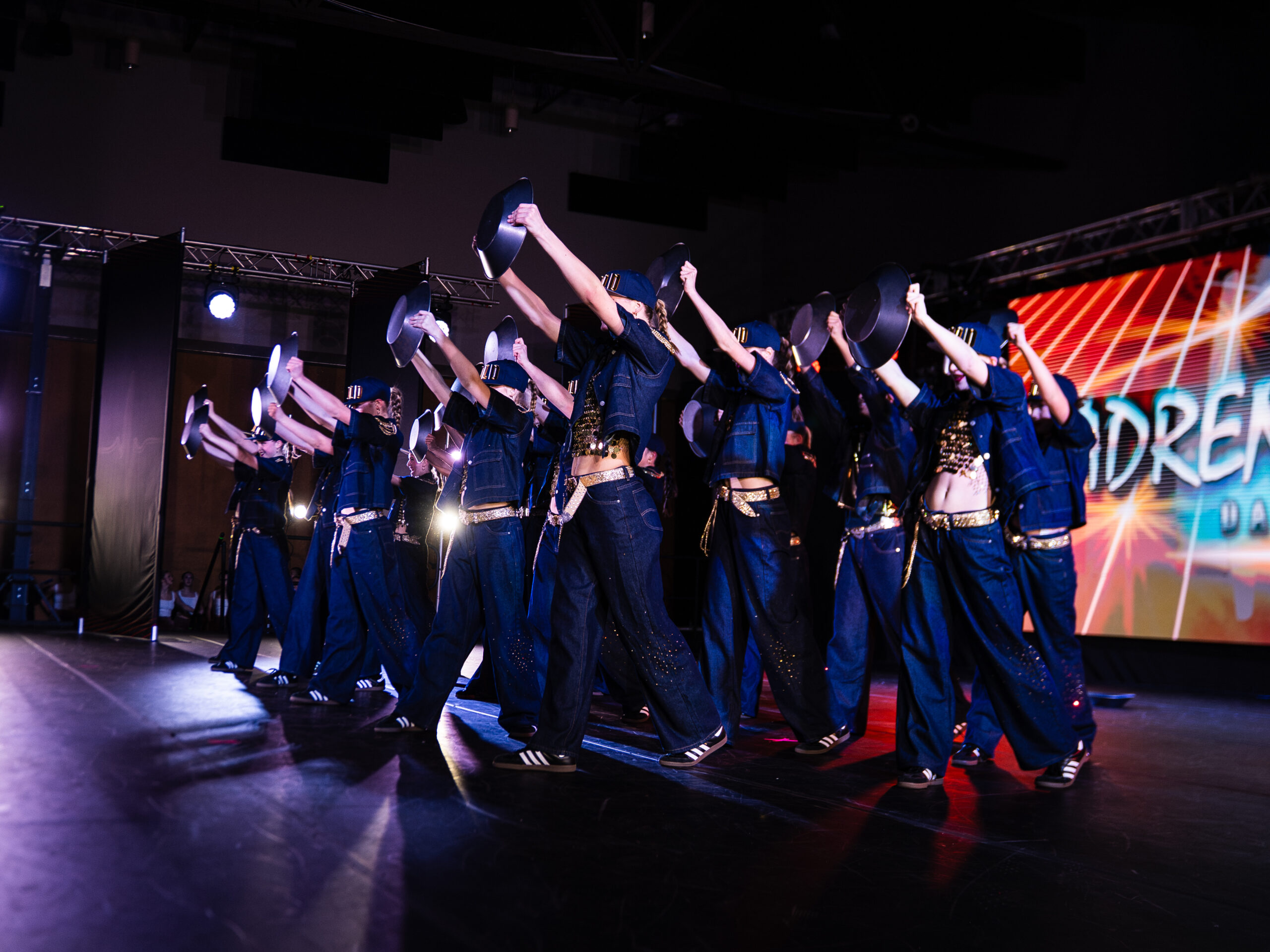 Dancers in blue outfits on stage, raising shiny metal plates overhead in a synchronized performance.