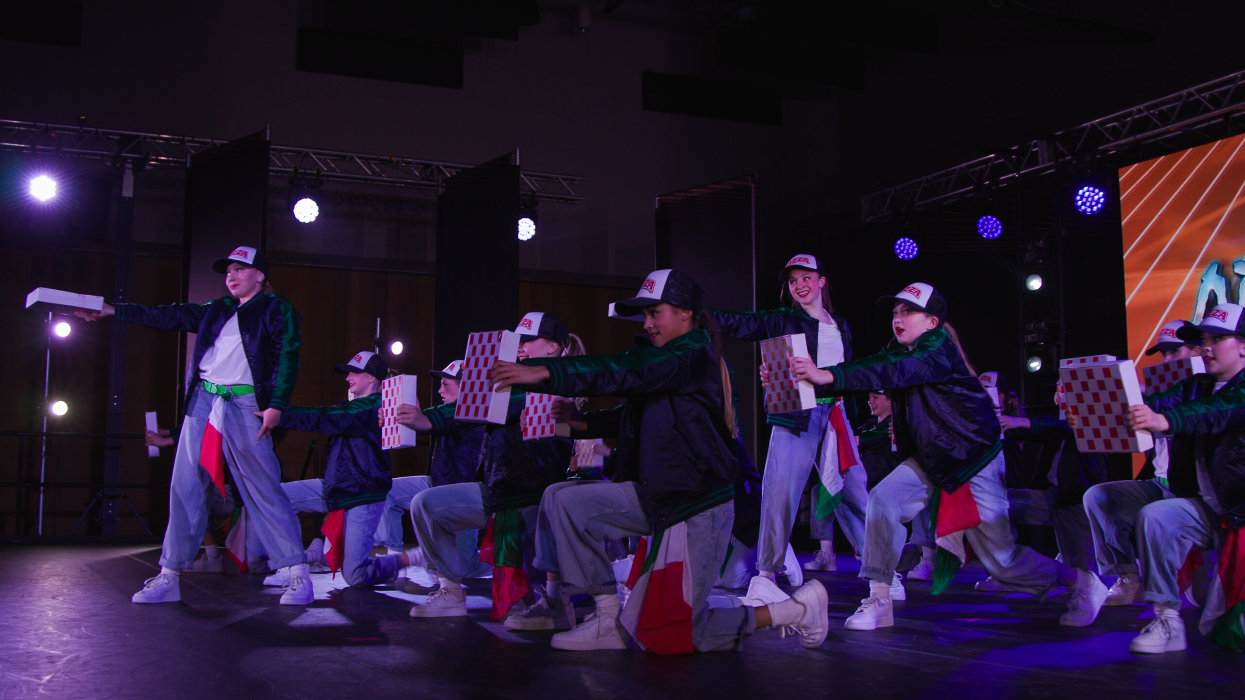 Dancers in green jackets and white caps perform a synchronized routine on a dark stage, holding white boxes with red checker patterns.