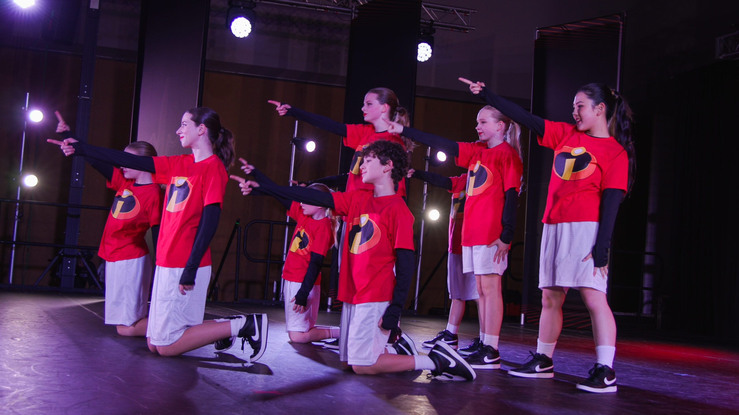 Group of young dancers in red shirts with black sleeves perform a choreographed pose on stage, pointing to the side.