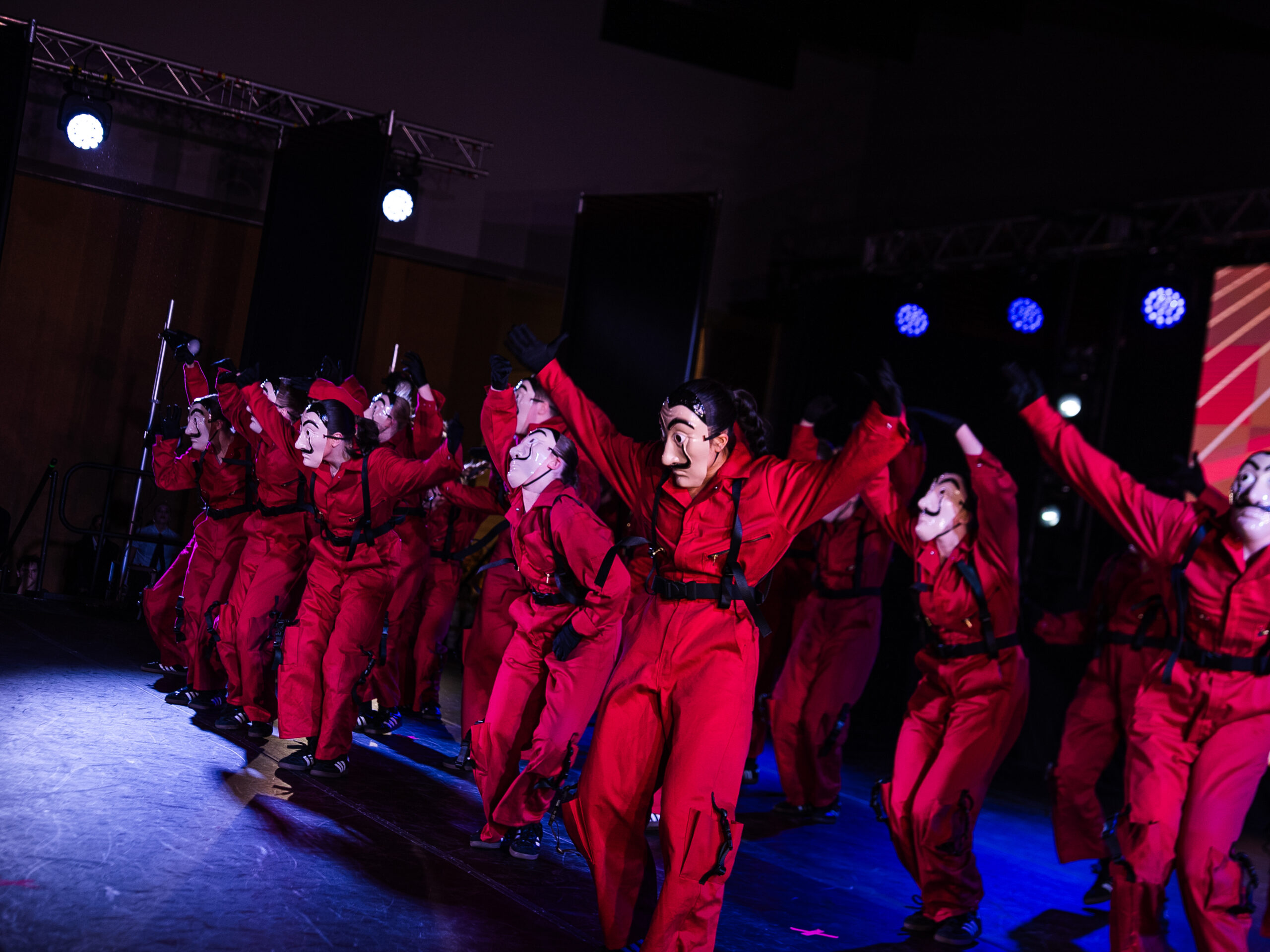 Group of performers in red jumpsuits and stylized masks dancing on a dimly lit stage with blue spotlights nearby