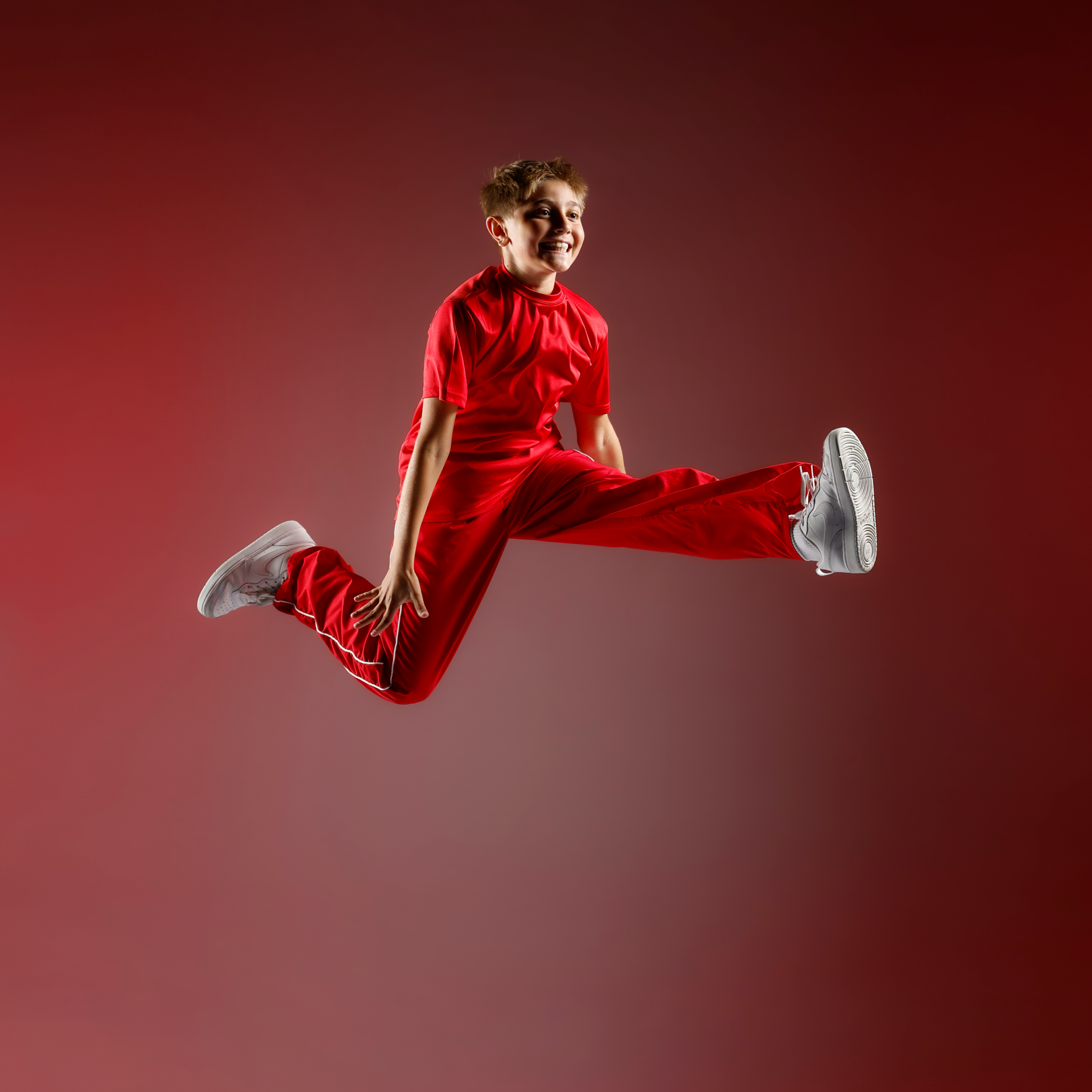 Boy in a red sports outfit jumping mid-air, white sneakers visible, against a red gradient background.