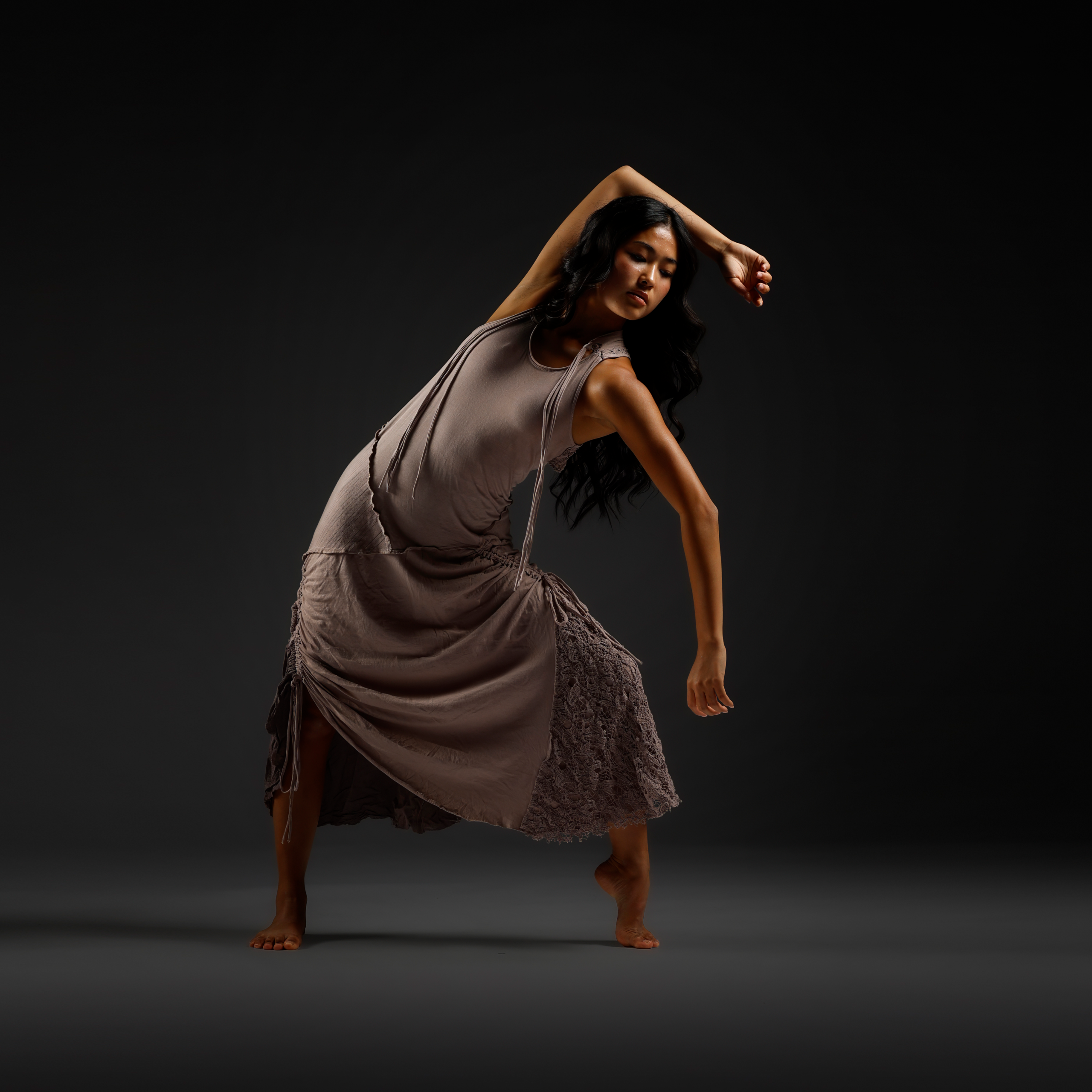 Contemporary dancer in a sleeveless beige dress, barefoot, arms raised in a graceful pose against a dark gradient background.