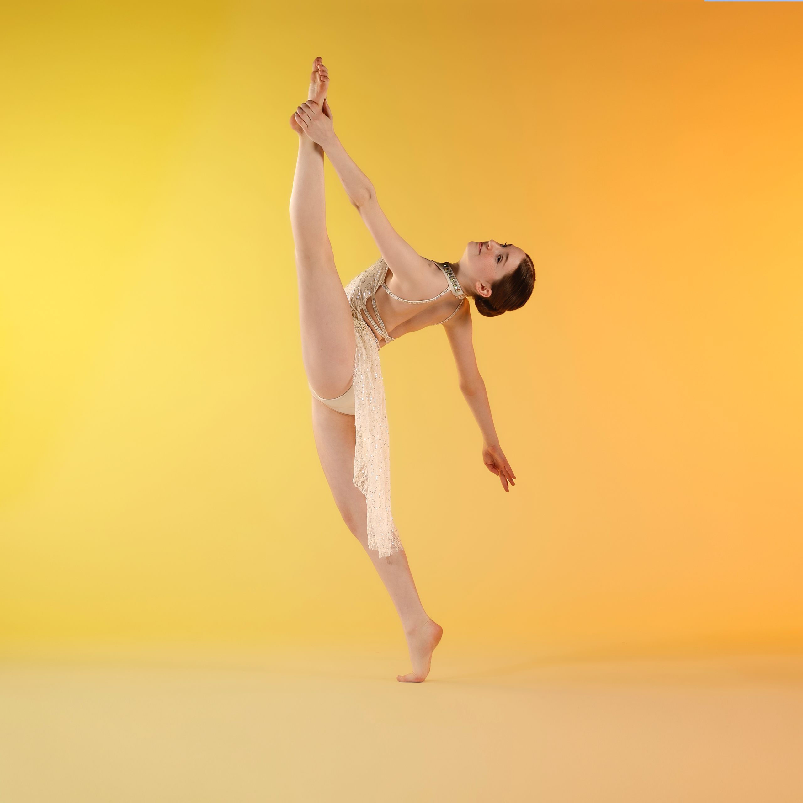 Dancer in a gold costume stretches a leg overhead, holding the ankle against a warm yellow‑orange gradient background.