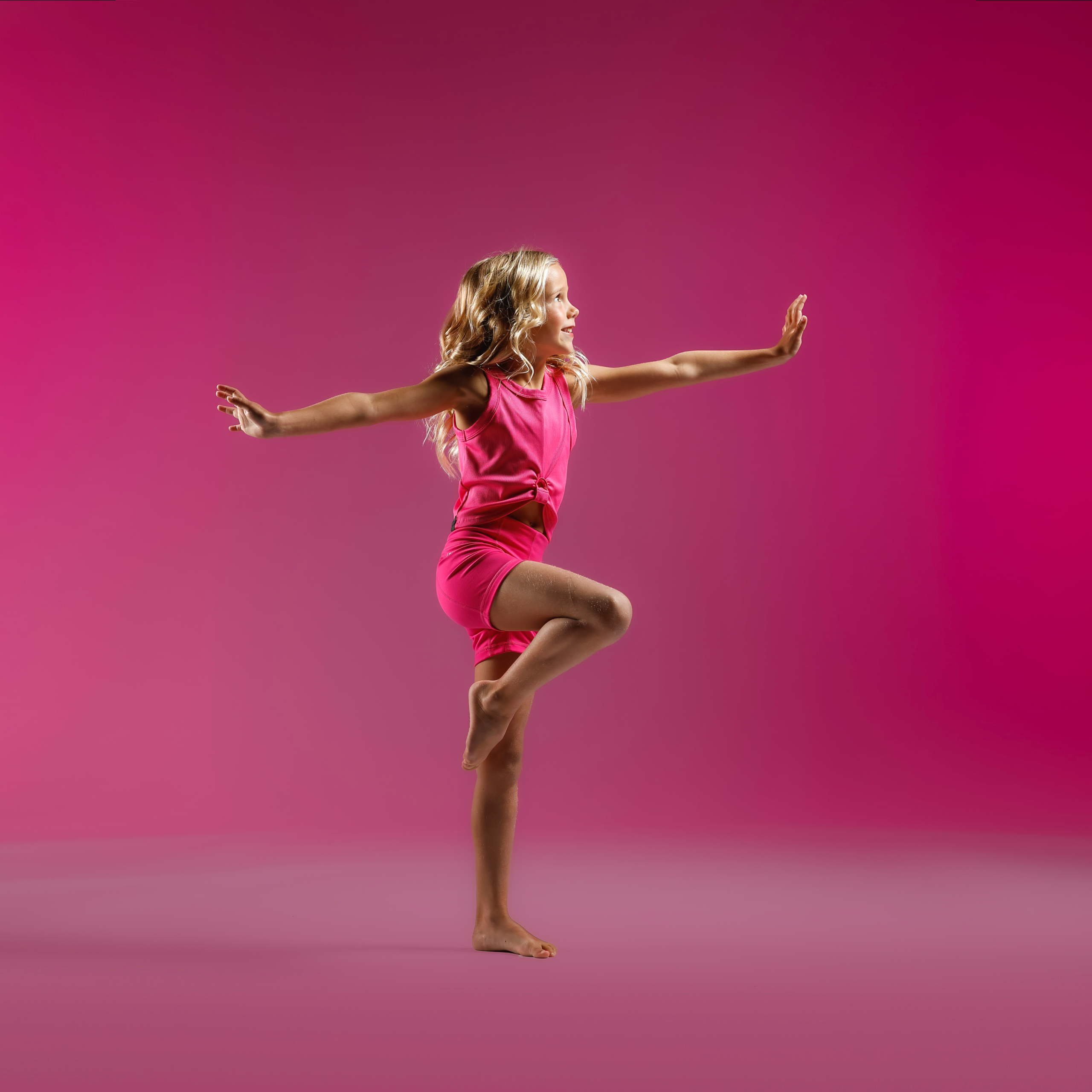 Girl in pink athletic outfit jumping mid‑air with arms extended against a pink gradient background, mid‑motion pose